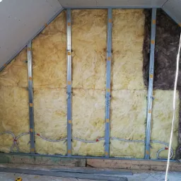 Skośny strych w trakcie izolacji: żółta wełna mineralna i ciemniejsza wełna w metalowym stelażu, widoczne przewody elektryczne i numeracja stelaża.