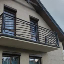 Czarna, metalowa balustrada balkonowa z poziomymi, zakrzywionymi prętami, zamontowana na balkonie domu z beżową elewacją i brązowymi ramami okiennymi.