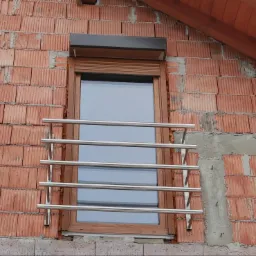 Nierdzewna balustrada okienna z poziomych rurek, zamontowana na elewacji z czerwonej cegły kratówki pod oknem z brązową ramą i roletą.