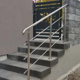 Zewnętrzne schody z ciemnych płyt z chromowaną balustradą, obok czarna brama z motywem roślinnym i mur z szarych bloków.