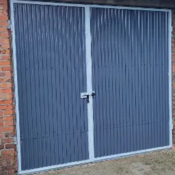 Dwuskrzydłowa brama garażowa z blachy falistej w szarej kolorystyce, osadzona w murze z czerwonej cegły, widoczny prosty zamek na środku bramy.
