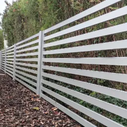 Nowoczesne ogrodzenie z poziomych, szarych profili, z widocznymi kroplami deszczu, na tle zielonych krzewów i drzew, z korą drzewną na ziemi.