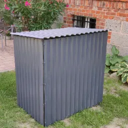 Szara, metalowa obudowa na licznik lub skrzynkę elektryczną, ustawiona na trawniku przed ceglanym budynkiem z kwiatami w tle.