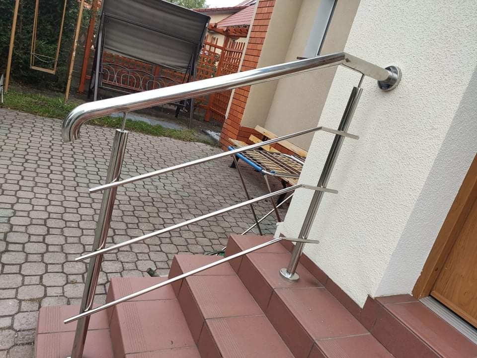 Zewnętrzna poręcz schodowa ze stali nierdzewnej, montowana do ściany budynku, widoczne płytki schodowe i kostka brukowa na podwórku.