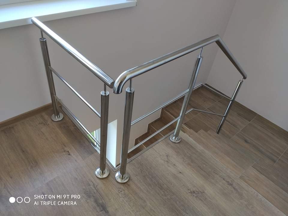 Polerowana, metalowa barierka schodowa z poziomymi poprzeczkami, zamontowana na schodach wyłożonych panelami imitującymi drewno.