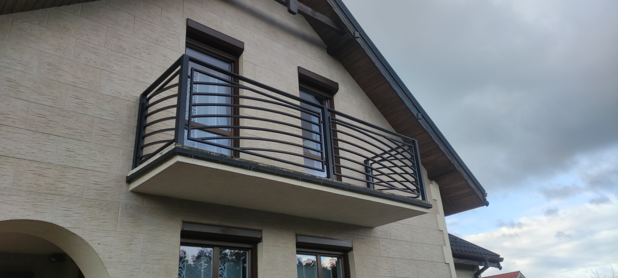 Czarna, metalowa balustrada balkonowa z poziomymi, zakrzywionymi prętami, zamontowana na balkonie domu z beżową elewacją i brązowymi ramami okiennymi.