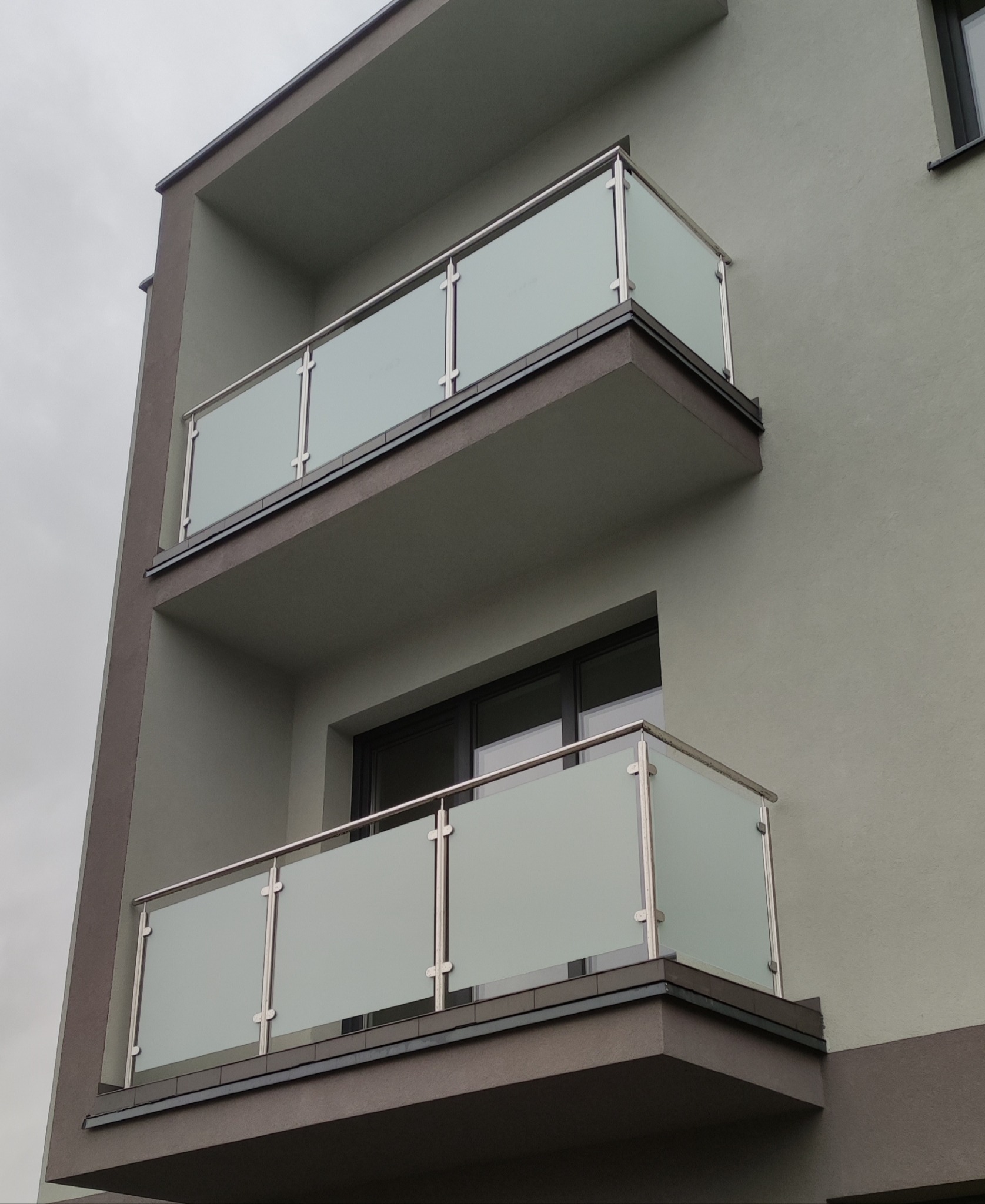 Dwa balkony z matowymi szklanymi panelami i nierdzewnymi balustradami, widok z dołu na tle pochmurnego nieba.