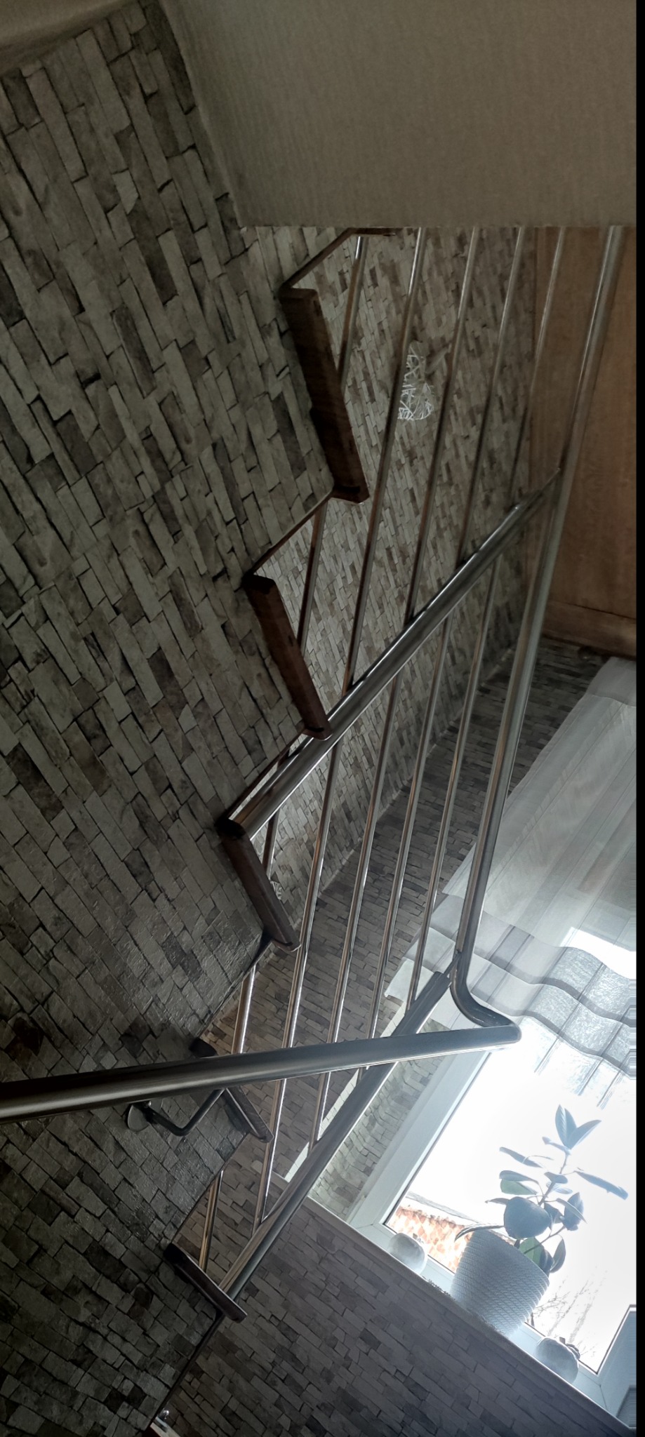 Wewnętrzne schody z metalową balustradą i drewnianymi stopniami, widok z góry, ściana wyłożona kamieniem dekoracyjnym, roślina doniczkowa przy oknie.