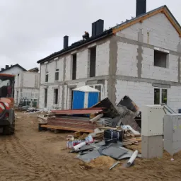 Wznoszenie dwukondygnacyjnego domu z białych bloczków, widoczny pracownik na dachu, pomarańczowy wózek widłowy w tle, teren budowy z materiałami i skrzynkami elektrycznymi.