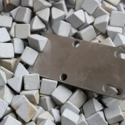 Metalowa płytka z otworami umieszczona wśród kostek ściernych o kształcie ostrosłupów i sześcianów, używanych w procesie obróbki wykończeniowej.