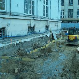 Usługi Inżyniersko - Budowlane Paweł Sobiech - Podkopywanie fundamentów starego budynku z ozdobnymi oknami, widoczny wykop, zabezpieczenia z żółtymi barierkami Skanska i mała koparka.