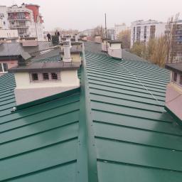 Usługi Inżyniersko - Budowlane Paweł Sobiech - Zielony blaszany dach z kominami na tle miejskiej zabudowy, dwóch mężczyzn na dachu w oddali.