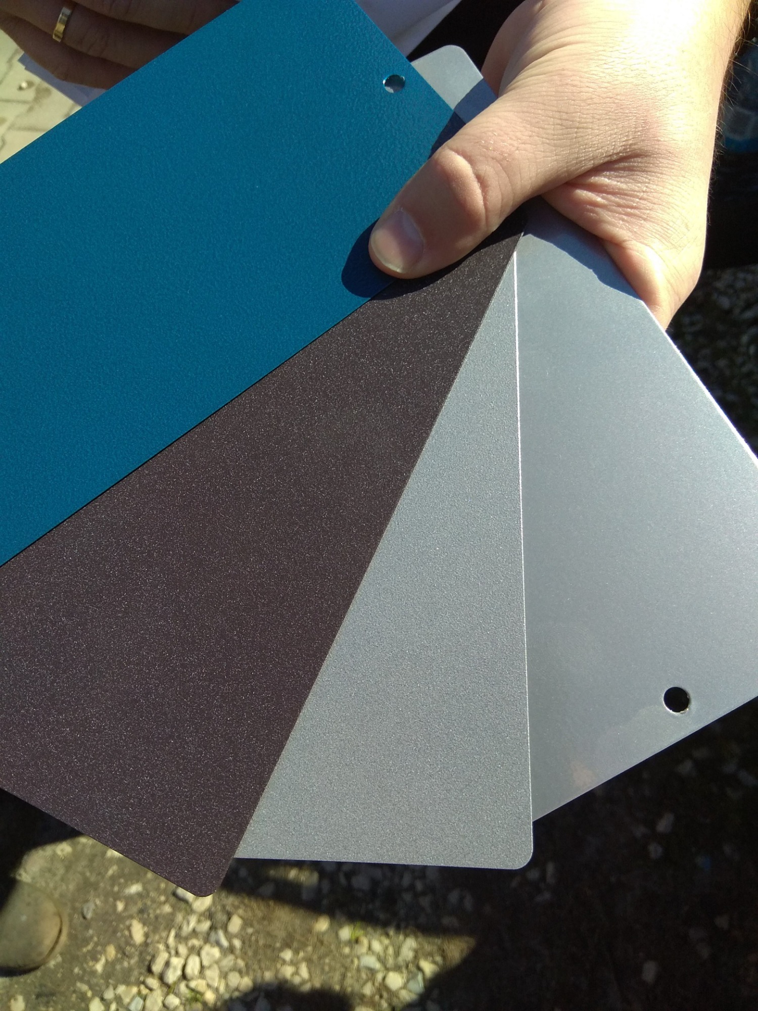 Dłoń trzymająca próbki kolorów metalu: turkusowy, grafitowy i srebrny, z widocznymi otworami montażowymi, prezentowane na zewnątrz.