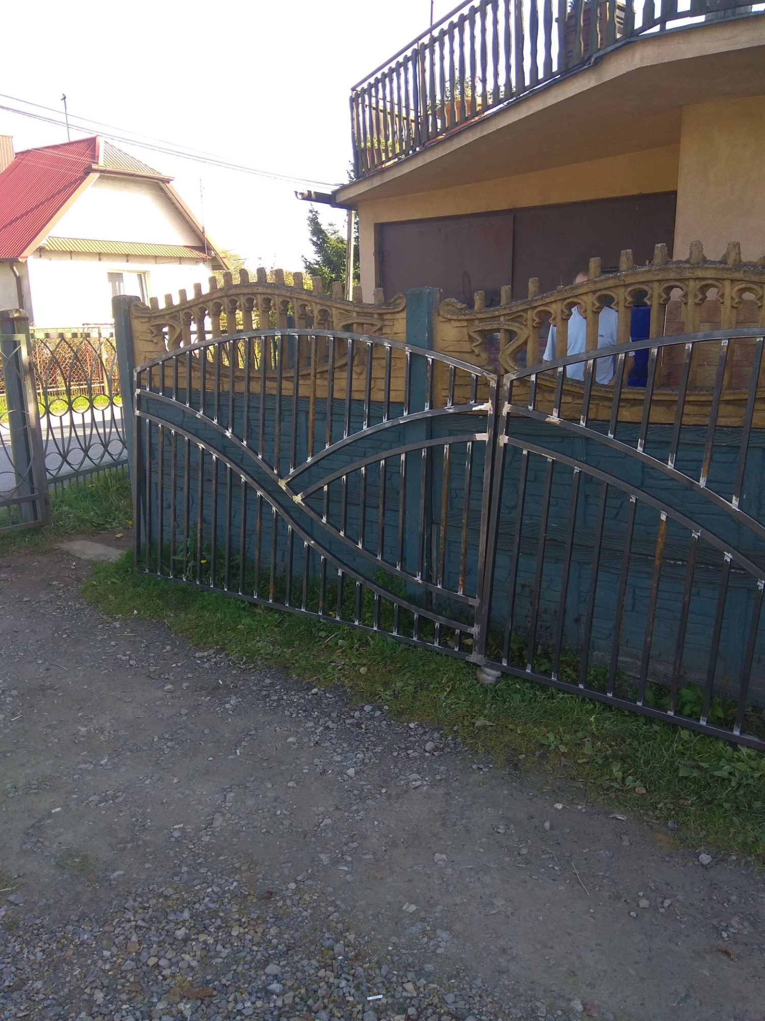 Ciemna, metalowa brama wjazdowa z łukowatym wzorem, widoczna z boku na tle zielonej ściany i fragmentu domu z balkonem. Na bramie widoczne ślady spawania.