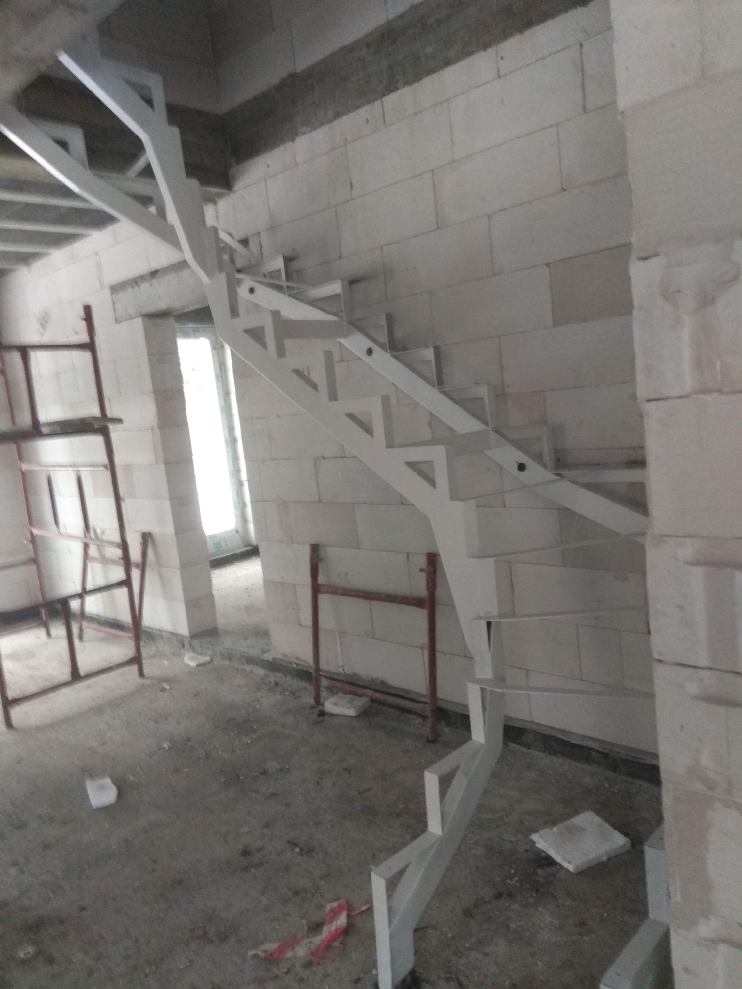 Surowe wnętrze w trakcie budowy z widocznymi schodami o metalowej konstrukcji, pomalowanej na biało, bez balustrady, przy ścianie z białych bloczków, z widocznym rusztowaniem w tle.