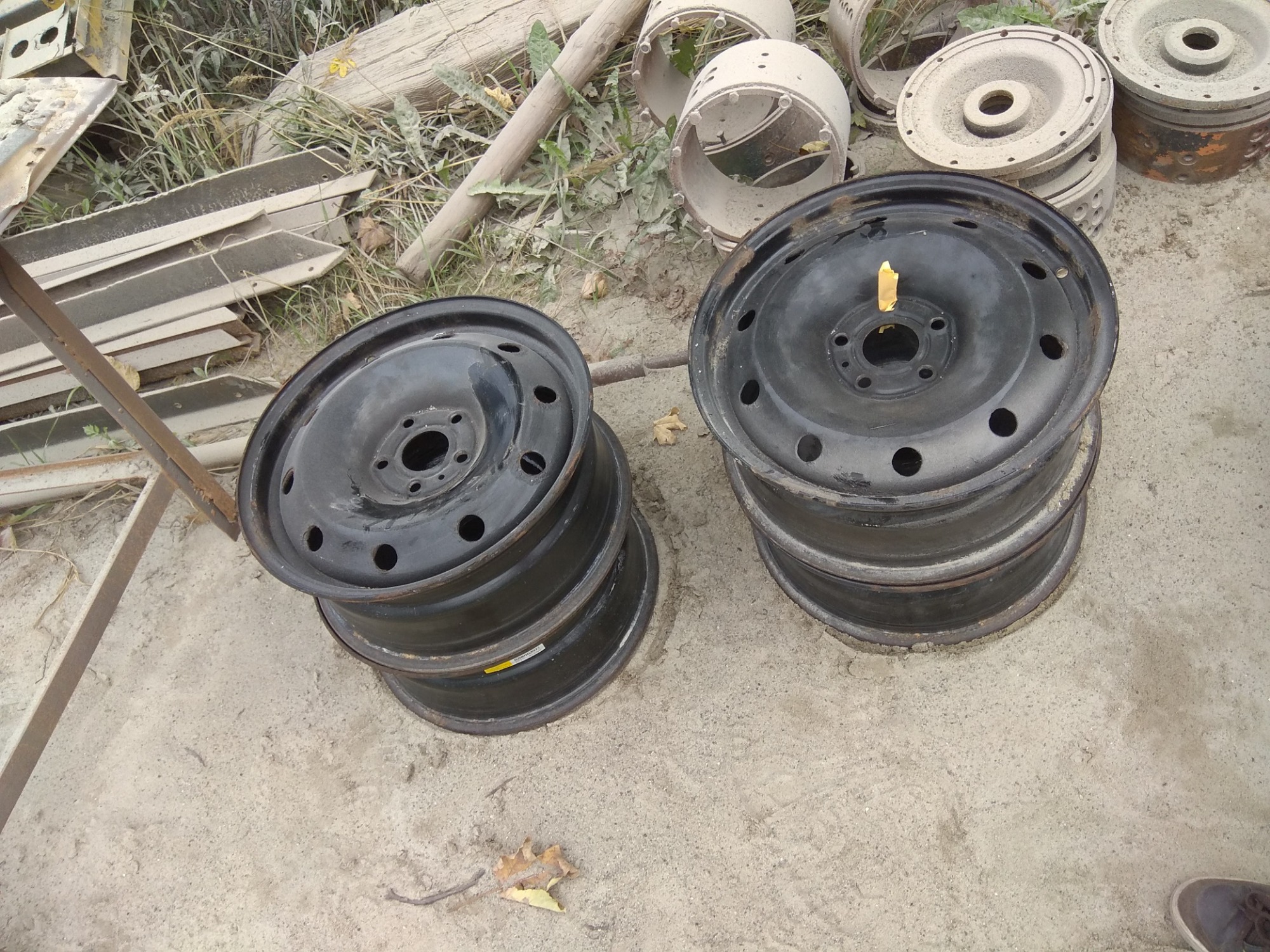 Dwa stosy czarnych, stalowych felg samochodowych na betonowym podłożu, otoczone metalowymi elementami konstrukcyjnymi i fragmentami drewnianych belek.