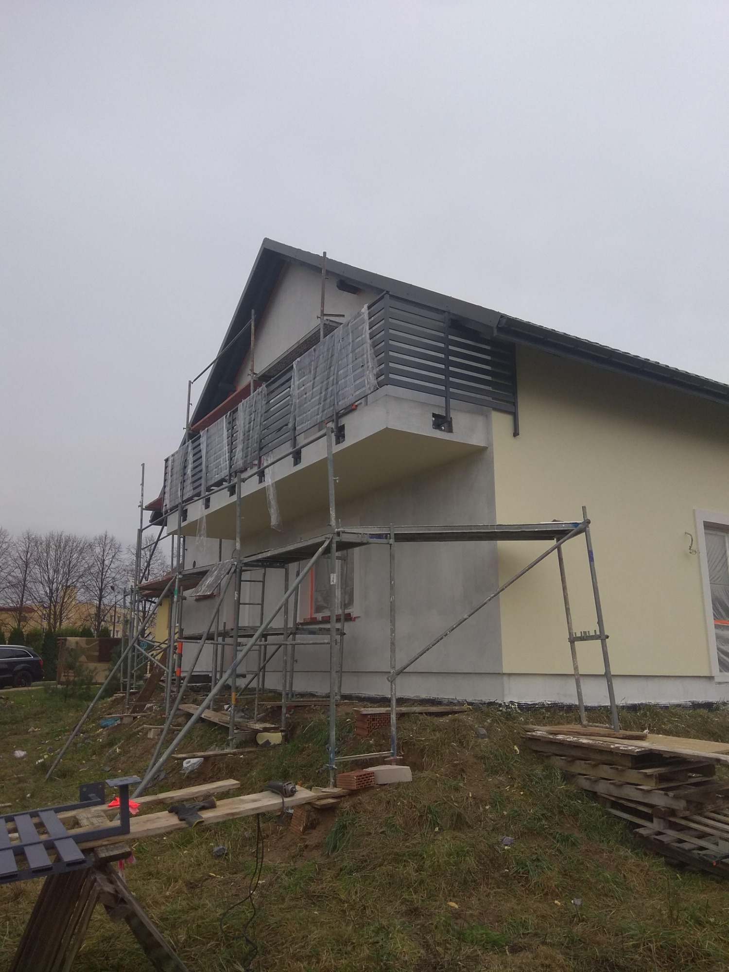 Montaż balustrady balkonowej z poziomych profili w Zaczerniu, widoczne rusztowania i zabezpieczenia folią, budynek w trakcie wykończenia elewacji.