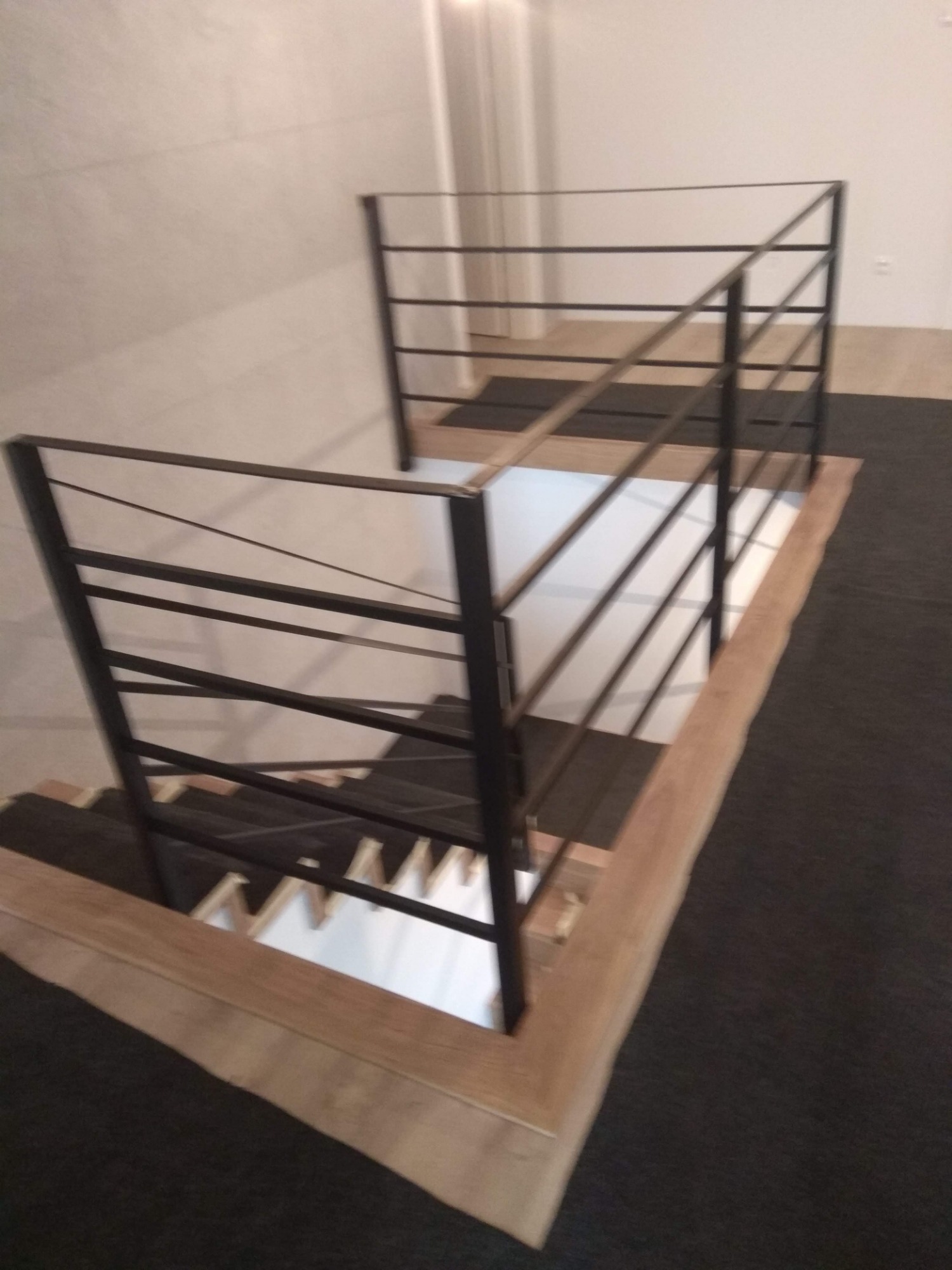 Wewnętrzna balustrada schodowa z czarnych, poziomych prętów, zamontowana na drewnianym podeście, widok z góry na schody wyłożone ciemną wykładziną.