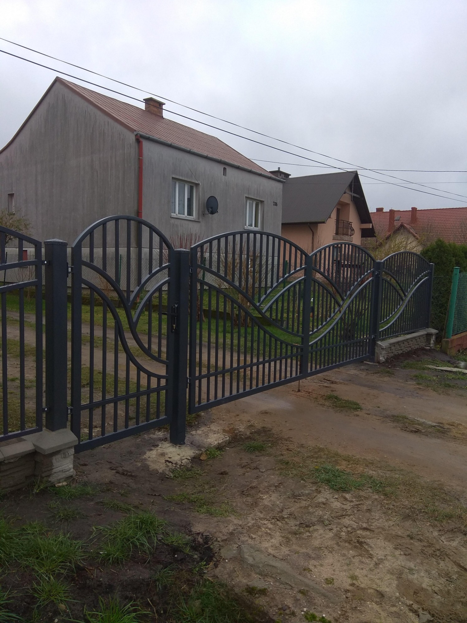 Szara, metalowa brama wjazdowa z ozdobnymi, łukowatymi elementami na tle szarego, dwupiętrowego domu z anteną satelitarną i pochmurnego nieba.