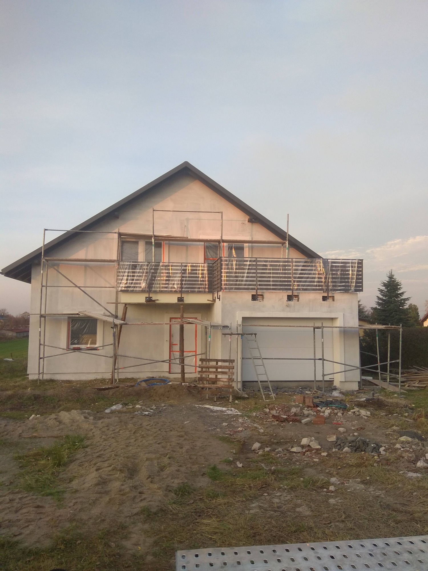 Dom w trakcie budowy z balkonem z balustradą nierdzewną, otoczony rusztowaniami i materiałami budowlanymi, widoczne cegły i drabina.