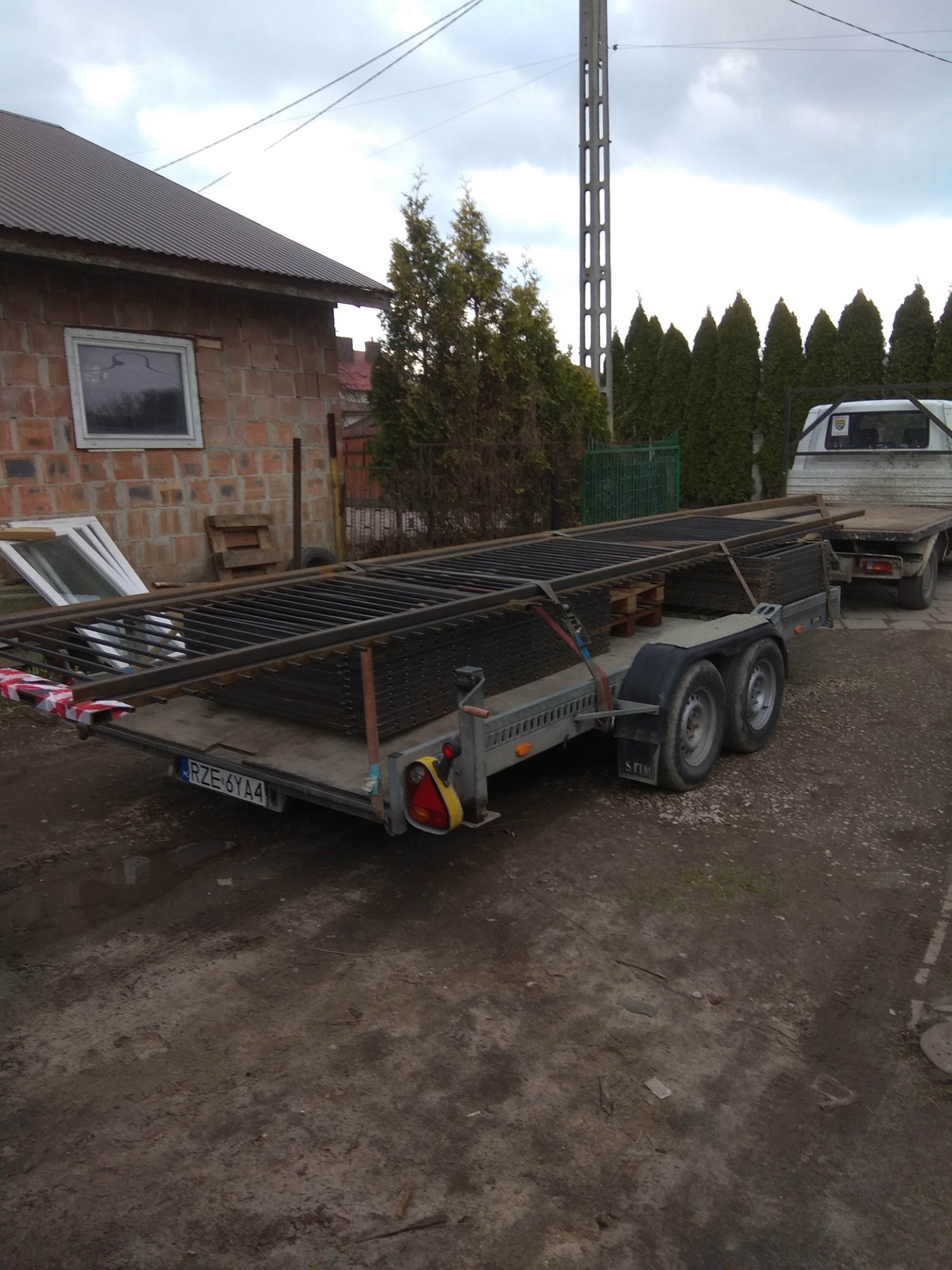 Przyczepa z ładunkiem czarnych, metalowych elementów ogrodzeniowych, zabezpieczonych pasami transportowymi, na tle domu z cegły i zielonych drzew.