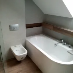 Nowoczesna łazienka na poddaszu z wanną, wiszącą toaletą myjącą i szarą glazurą, drewnopodobne akcenty