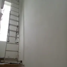 Świeżo pomalowana, jasna ściana w trakcie prac wykończeniowych, z aluminiową drabiną opartą o ścianę przy oknie i zabezpieczonymi schodami na pierwszym planie.