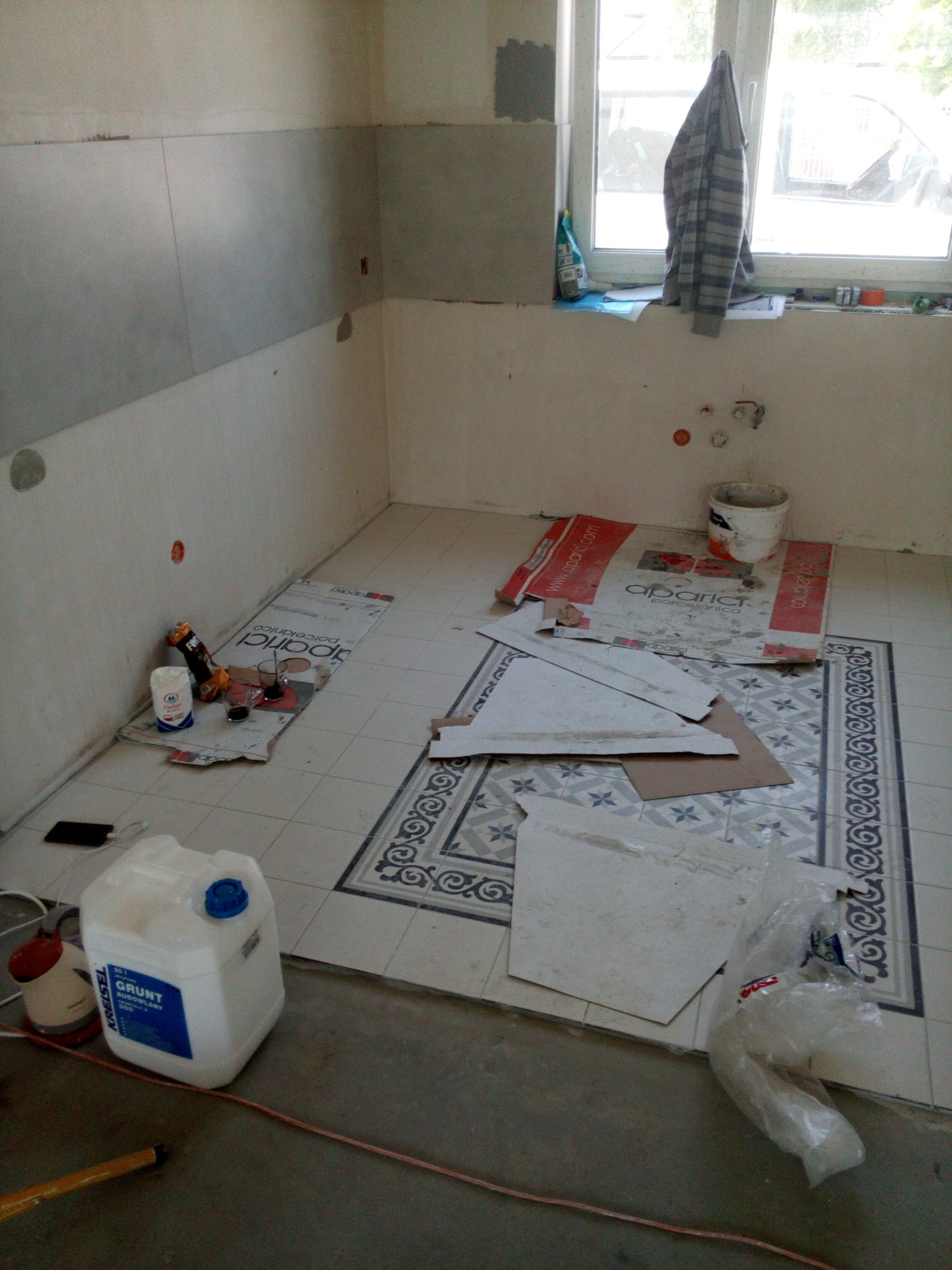 Fragment wnętrza w trakcie prac wykończeniowych: częściowo położone płytki z dekoracyjnym wzorem na podłodze, widoczne narzędzia, grunt budowlany i resztki materiałów budowlanych, ściany częściowo...