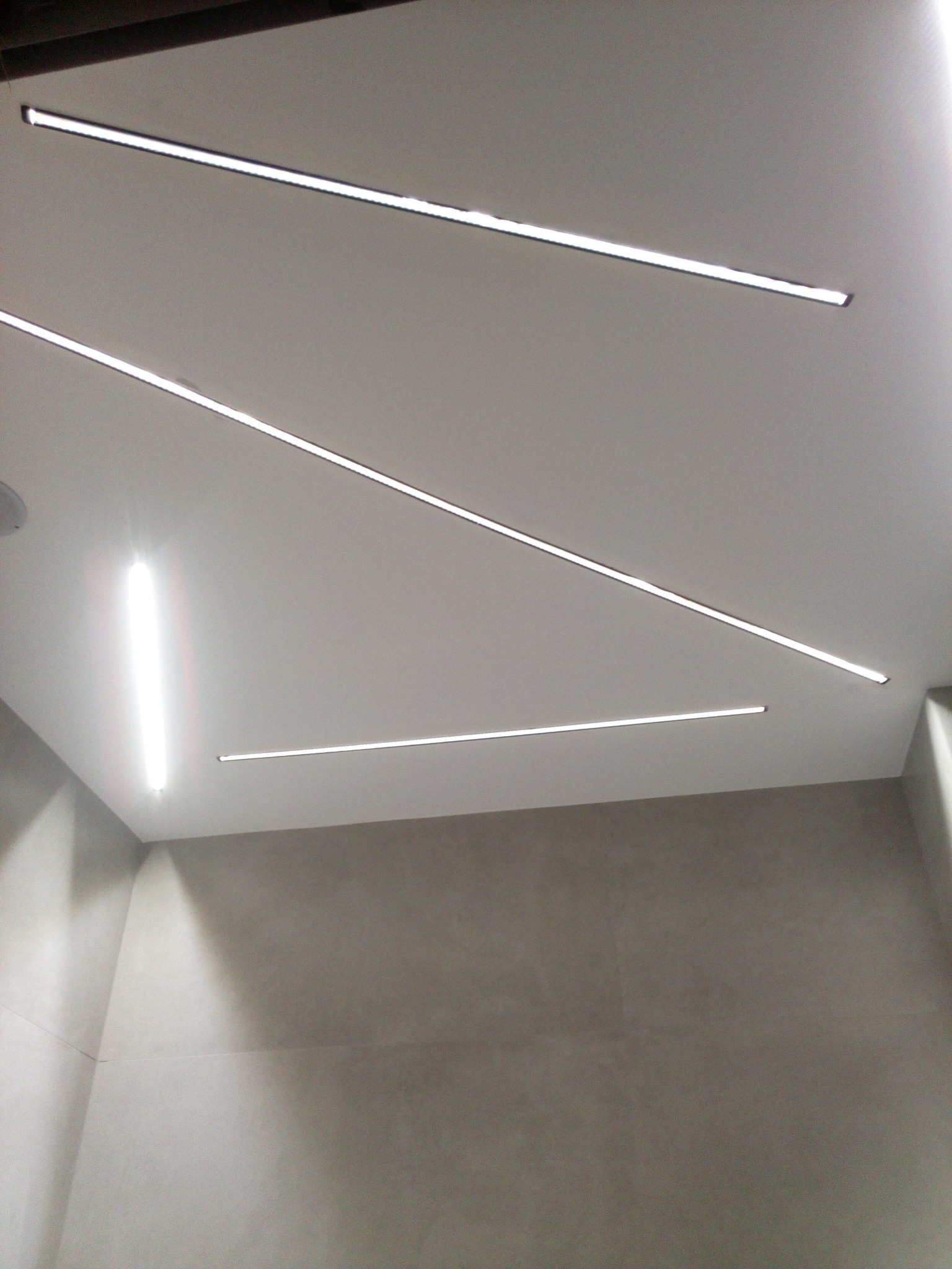 Nowoczesne oświetlenie łazienkowe: trzy linearne lampy LED wpuszczone w sufit, szare płytki na ścianach.