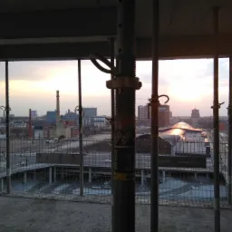 Widok z surowego budynku w trakcie budowy na miasto o zachodzie słońca, przez metalowe podpory stropowe i siatkę zabezpieczającą.