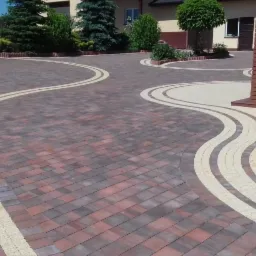 Wzorzysta nawierzchnia z kostki brukowej w odcieniach brązu i beżu, ułożona w falujące linie, prowadząca do wejścia domu z ceglanymi filarami.