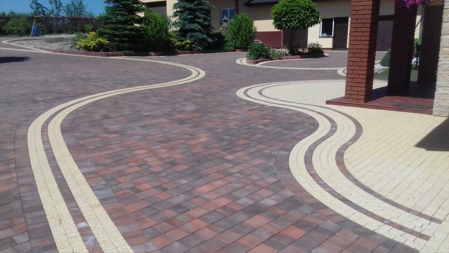 Wzorzysta nawierzchnia z kostki brukowej w odcieniach brązu i beżu, ułożona w falujące linie, prowadząca do wejścia domu z ceglanymi filarami.