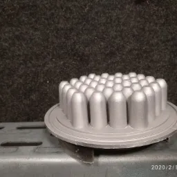 Metalowy element z wypustkami po obróbce, leżący na metalowej półce na tle ciemnej, teksturowanej ściany.