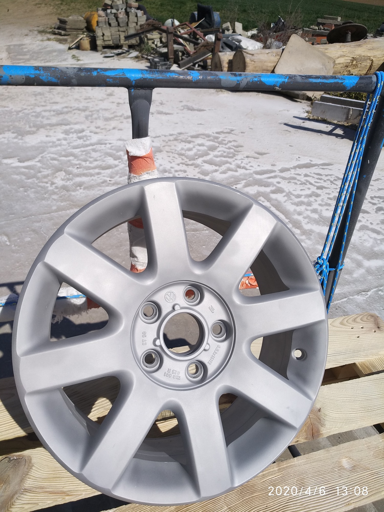 Pomalowana na szaro felga samochodowa Volkswagen, zawieszona na metalowym stojaku z niebieskimi elementami, na tle pobielonej powierzchni i drewnianej palety.