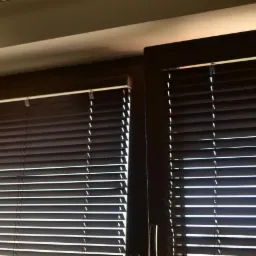 Ciemnobrązowe drewniane żaluzje poziome w oknie, z widocznymi sznurkami i listwami, częściowo zasłonięte, z fragmentem białej zasłony po lewej stronie.