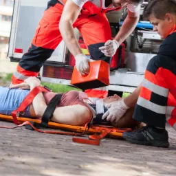 Ratownicy medyczni udzielają pomocy kobiecie leżącej na noszach na chodniku z cegły, w tle ambulans i budynek.