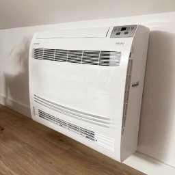 Solar Wójcik sp. z o.o. - Biały klimatyzator podłogowy marki Aneru, zamontowany przy ścianie na drewnianej podłodze, widok z boku.