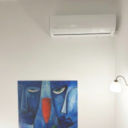 Solar Wójcik sp. z o.o. - Biały klimatyzator na ścianie nad abstrakcyjnym obrazem w odcieniach błękitu i czerwieni, oświetlony ciepłym światłem lampki.