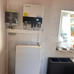 Solar Wójcik sp. z o.o. - Instalacja fotowoltaiczna w pomieszczeniu: inwerter Growatt, skrzynka z bezpiecznikami i akumulator. Widoczne okablowanie i elementy montażowe.