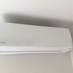 Solar Wójcik sp. z o.o. - Biała jednostka klimatyzacji Rotenso zamontowana na ścianie w jasnym pomieszczeniu. Widoczny fragment sufitu i ściany, urządzenie w centralnej części kadru.