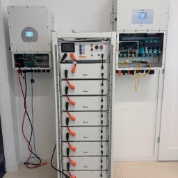 Solar Wójcik sp. z o.o. - Biała szafa z modułami bateryjnymi Deye, zasilana z dwóch białych inwerterów na ścianie. Widoczne okablowanie i podłączenia. Całość w jasnym, minimalistycznym wnętrzu.