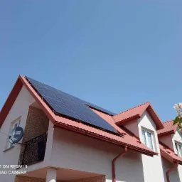 Dom z czerwonym dachem i panelami słonecznymi w słoneczny dzień. Widoczny balkon z ozdobną balustradą i satelitą. W tle błękitne niebo i kwitnące drzewo.