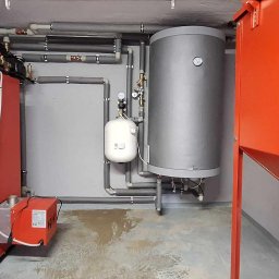 Solar Wójcik sp. z o.o. - Czerwony piec marki Rakoczy Firemax z osprzętem, zbiornik buforowy i instalacja grzewcza z szarych rur w kotłowni, widok na całość instalacji.