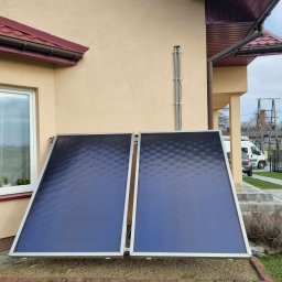 Solar Wójcik sp. z o.o. - Dwa płaskie kolektory słoneczne zamontowane przy ścianie domu z beżową elewacją, widoczne okno i fragment dachu. Tło stanowi trawnik i fragment ulicy.