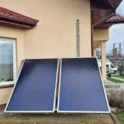 Dwa płaskie kolektory słoneczne zamontowane przy ścianie domu z beżową elewacją, widoczne okno i fragment dachu. Tło stanowi trawnik i fragment ulicy.