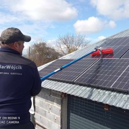 Solar Wójcik sp. z o.o. - Mężczyzna w uniformie SolarWójcik czyści czerwoną szczotką panele fotowoltaiczne na blaszanym dachu. W tle błękitne niebo i drzewa.