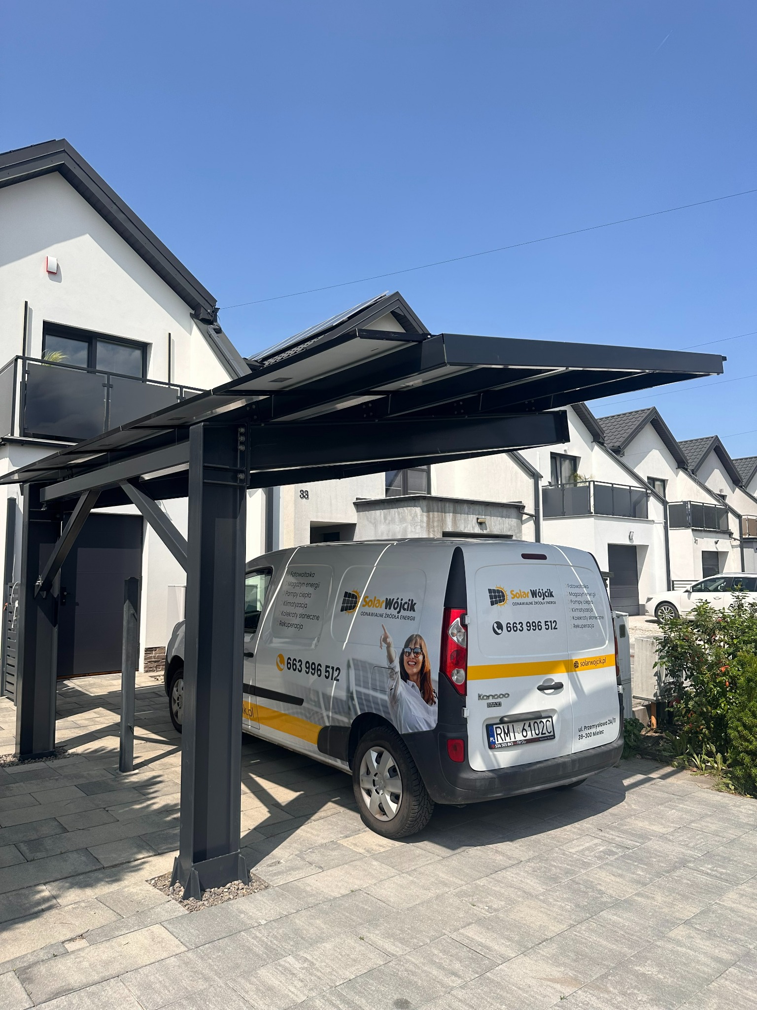 Carport z panelami fotowoltaicznymi i zaparkowany pod nim biały bus firmy Solar Wójcik na tle nowoczesnych domów jednorodzinnych w słoneczny dzień.