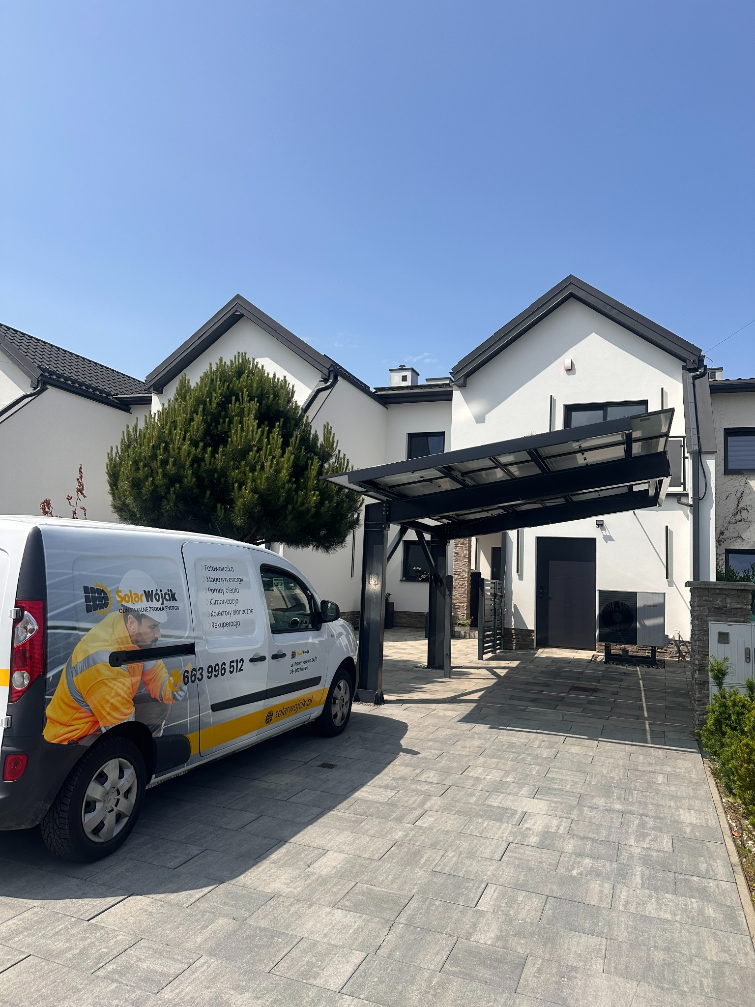 Biały dom z czarną stolarką i zadaszeniem fotowoltaicznym. Na podjeździe stoi firmowy bus Solar Wójcik z reklamą fotowoltaiki i pomp ciepła.