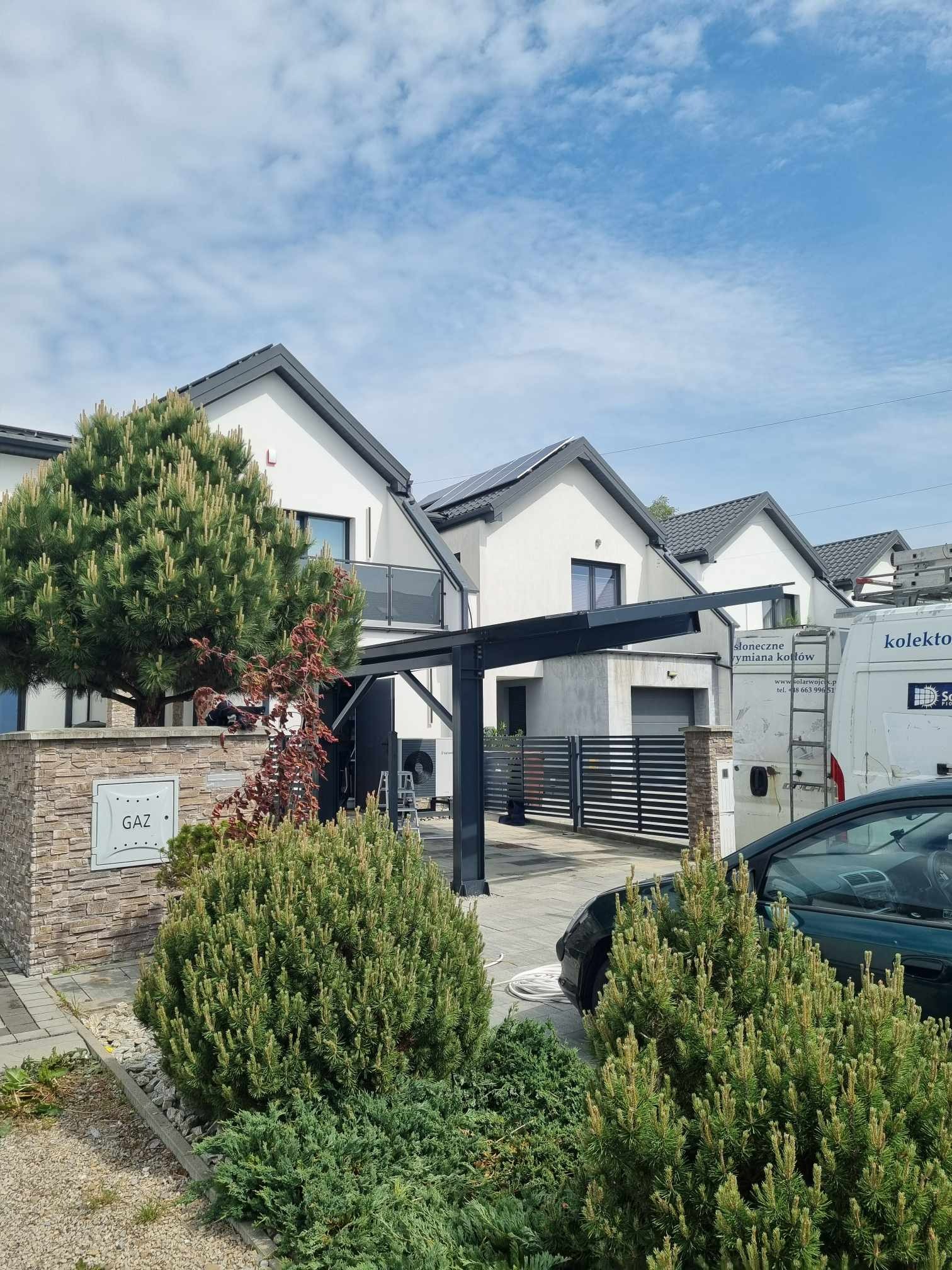 Nowoczesne domy z panelami słonecznymi na dachu, widoczne spod zadaszenia garażowego. Na pierwszym planie zieleń i skrzynka gazowa.