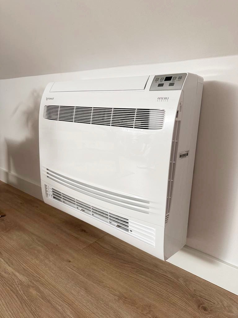 Biały klimatyzator podłogowy marki Aneru, zamontowany przy ścianie na drewnianej podłodze, widok z boku.